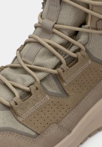 Bota de caminhada castanha com um upper de tecido texturizado, ilhós reforçados e cordões elásticos. Apresenta acabamentos em camurça castanha e buracos de ventilação.