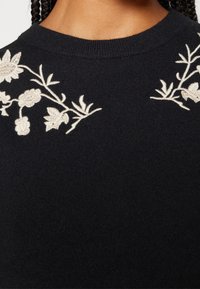 Pull-over bleu marine foncé avec une broderie florale crème complexe le long de l'encolure. Texture tricotée, col rond, coupe ajustée.