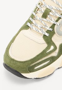 Beige hálós felsőrésszel rendelkező sportcipők, ol�íva zöld bársony részletekkel, texturált krém színű középtalppal, valamint mintás fehér és fekete fűzőkkel.