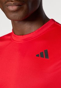 Camiseta deportiva roja con una textura suave, que presenta un logo negro en la parte superior izquierda. El cuello es redondo y ajustado.