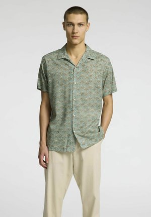 Camisa de manga corta en tejido azul claro con un patrón repetido de hojas doradas, con un corte relajado y cuello puntiagudo. Combinándola con pantalones beige.