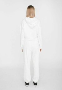 Vitt set med hoodie och matchande mjukisbyxor, tillverkat av mjukt tyg. Har huva med dragsko och elastiska muddar. Bärs med svarta sneakers.