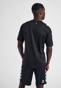 Zwarte sporttrui met korte mouwen, gemaakt van een gladde stof. Bevat witte chevronstrepen op de schouders en een logo aan de onderrand.