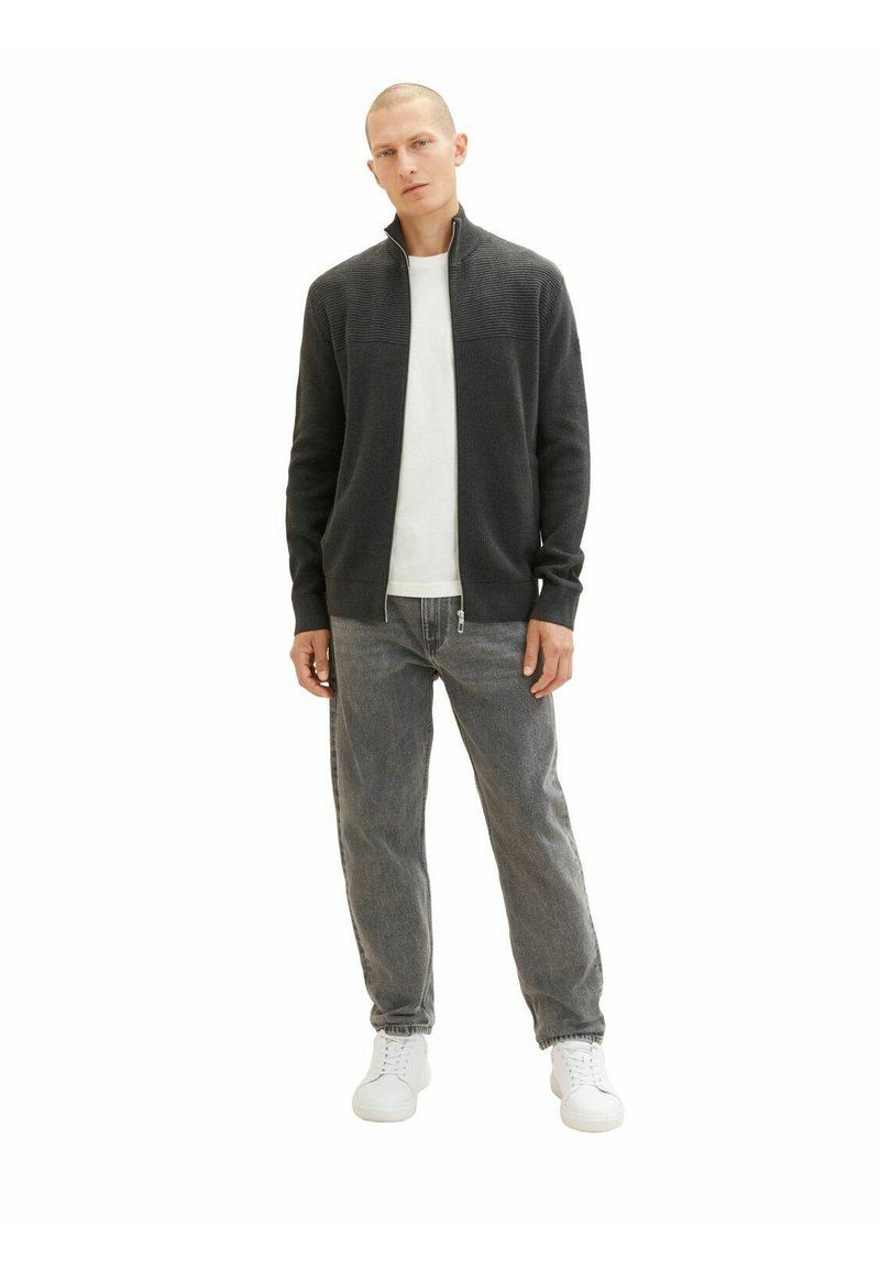 Szary kurtka zapinana na zamek z wysokim kołnierzem, noszona na białej koszulce z okrągłym dekoltem. Szare dżinsy i białe sneakersy dopełniają strój. Gładka faktura.