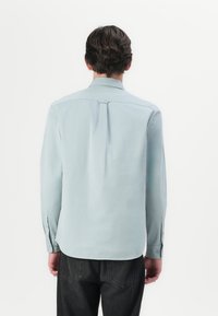 Camisa de manga longa azul claro com uma textura suave, colarinho padrão e botões nos punhos. As costas apresentam um detalhe de costura vertical.