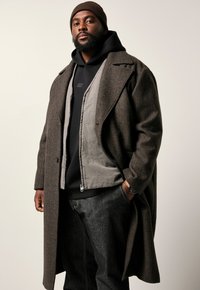 Brauner Fischgräten-Mantel über einer grauen Zip-Jacke, getragen mit einem schwarzen Hoodie und dunklen Jeans. Dunkle Strickmütze auf dem Kopf.
