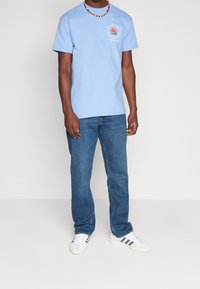 Světle modré bavlněné tričko s kulatým výstřihem a grafikou na hrudi. V kombinaci se středně modrými džíny a bílými teniskami s černými pruhy.