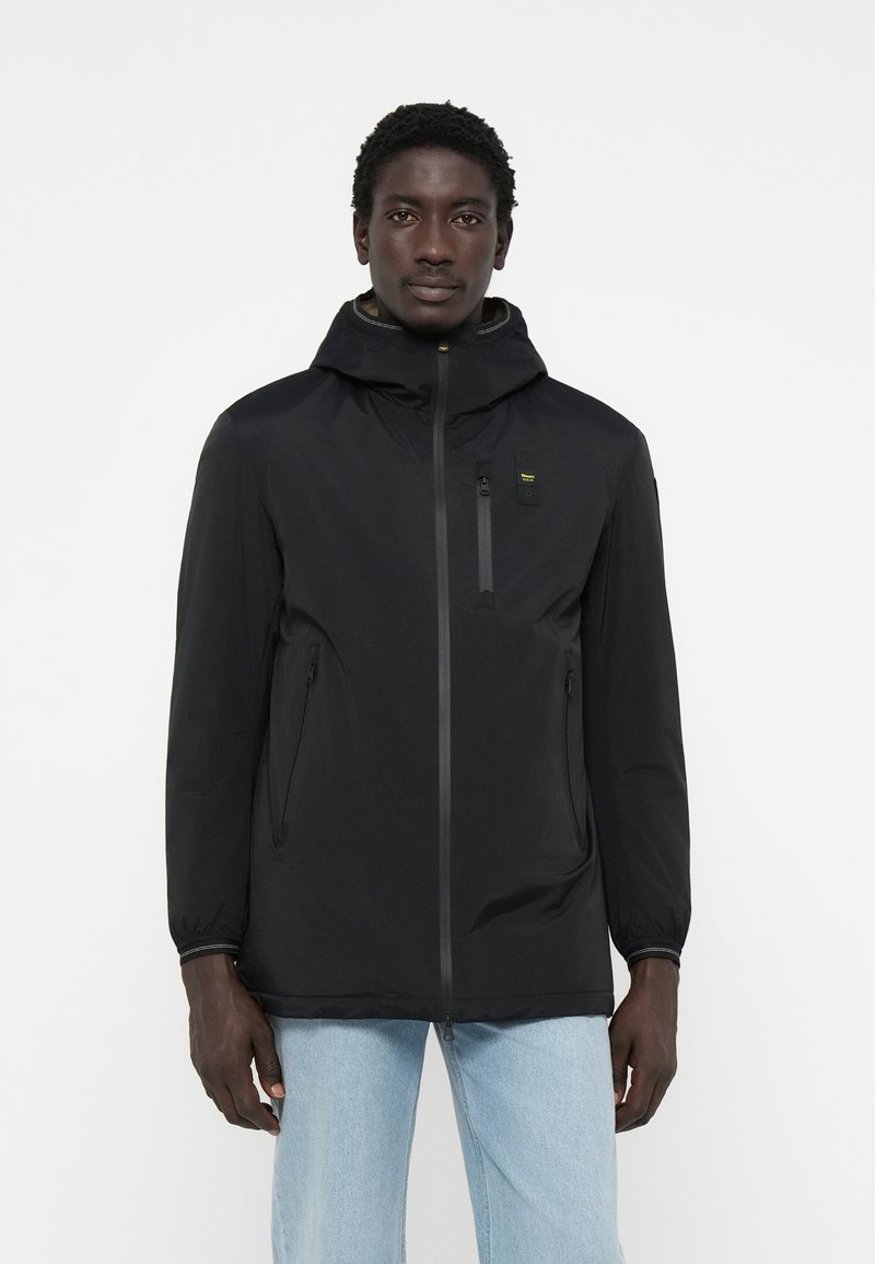 Schwarze leichte Jacke mit Kapuze, ausgestattet mit einem Frontreißverschluss, zwei Seitentaschen und einer Brusttasche mit gelben Akzenten. Glatte Textur.