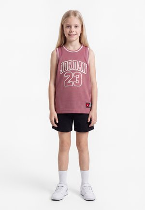 Jeune fille debout et souriante, portant un maillot de sport sans manches Jordan 23 rose, un short noir, des chaussettes blanches et des baskets blanches.