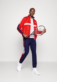 Chaqueta deportiva roja con rayas blancas, pantalones azul marino con acentos laterales y zapatillas deportivas blancas. El modelo sostiene una raqueta de tenis.