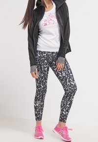 Musta vetoketjullinen takki, valkoinen t-paita, jossa vaaleanpunainen logo, kuviolliset mustat legginsit ja vaaleanpunaiset urheilukengät. Takissa on harmaat hihansuut.