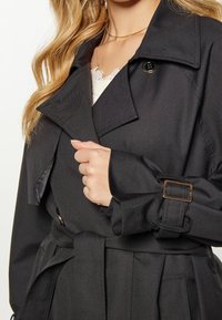 Trench-coat noir avec un large col, fermeture avant à boutons, taille ceinturée et tissu texturé. Comprend des accents de poignets avec une boucle en métal.