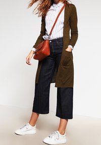 Długi kardigan oliwkowozielony na niebiesko-pasiastą koszulę z guzikami, w połączeniu z wysokim stanem ciemnych dżinsowych culottes i białymi trampkami, trzymający brązową torbę.