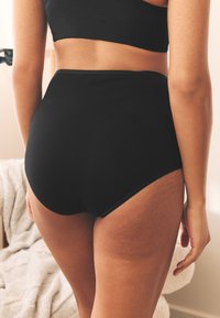 Schwarze Unterwäsche mit hoher Taille, glatter Textur und gewellten Kanten, vor hellem Hintergrund positioniert. Material aus Baumwollmischung.