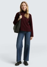 Bordeauxroter Rollkragenpullover kombiniert mit blauen Jeans mit weitem Bein und schwarzen Plateau-Stiefeln; Modell hält eine große schwarze Tote-Tasche.