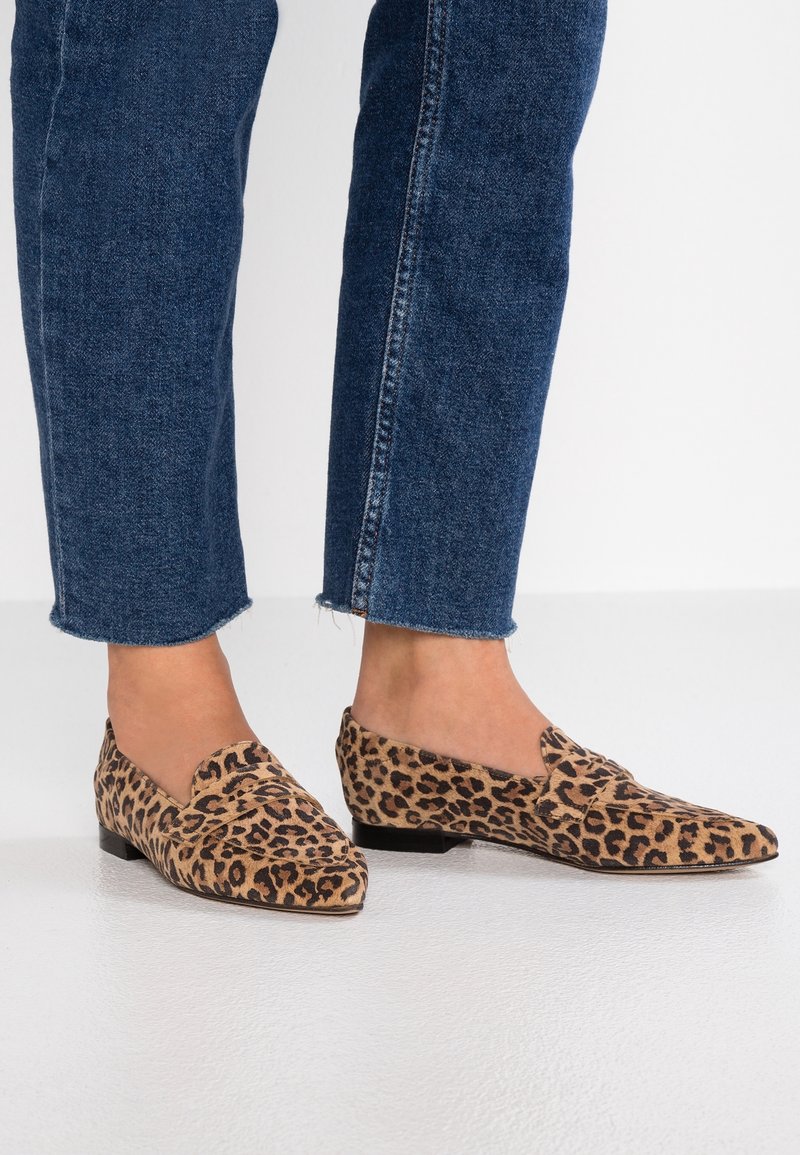 Des pieds chaussés de mocassins à imprimé léopard, associés à un jean en denim bleu foncé coupé, se tenant sur un sol blanc.