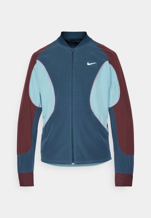 Chaqueta de pista de Nike con cierre frontal de cremallera, paneles azul y marrón, puños acanalados y un pequeño logo blanco en el pecho. Textura de tela suave.