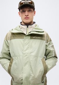 Light green hooded jacket with tan accents, zip front, and two front pockets. Features breathable mesh details and Columbia logo on chest.