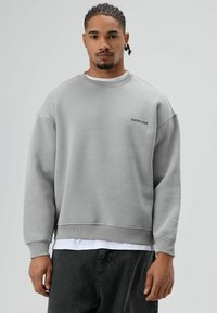 Grijze sweatshirt met doorgezakte schouders, geribbelde manchetten en een cropped zoom. Voorzien van geborduurde tekst "RAÍCES VIVAS" op de borst. Witte onderlaag zichtbaar.