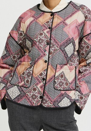 Veste matelassée avec motif patchwork rose, beige et noir, boutons noirs et poche avant, portée sur un top blanc et un pantalon gris foncé.