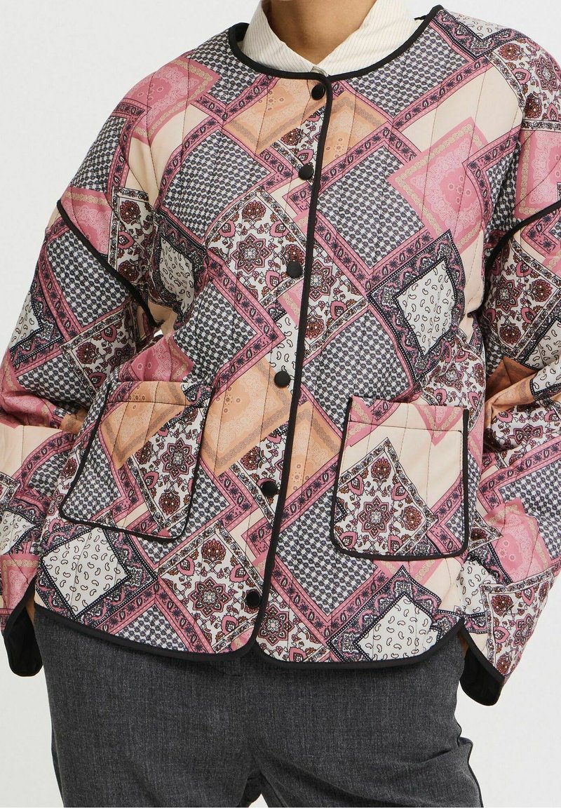 Veste matelassée avec motif patchwork rose, beige et noir, boutons noirs et poche avant, portée sur un top blanc et un pantalon gris foncé.