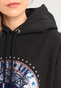 Musta huppari, jossa on suuri pyöreä paljetti kuvio, joka sisältää sinisiä, punaisia ja hopeisia elementtejä. Hupparin reunassa on kiristysnapit ja hopeiset reikäkohdat.