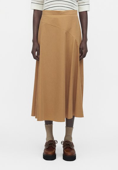 Femme portant une jupe midi beige, des chaussettes beiges, des chaussures marron à lacets et un haut blanc à motifs avec des manches jusqu'aux coudes.