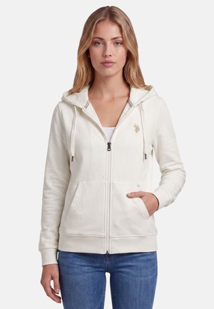 Woman wearing a white zip-up hoodie with a small logo on the chest, blue jeans, standing with one hand in pocket.