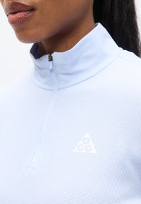 Ανοιχτό μπλε τοπ με τέταρτο φερμουάρ, κατασκευασμένο από υφασμάτινη υφή, με μικρό λευκό τριγωνικό λογότυπο στο στήθος και σχέδιο κολάρου με φερμουάρ.