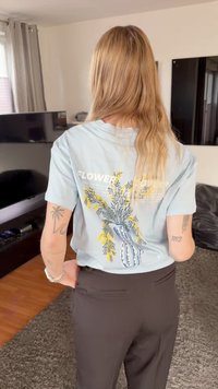 Mujer rubia con cabello largo está de pie en el interior, lleva una camiseta azul con un diseño de jarrón amarillo con flores y pantalones negros, manos en las caderas.