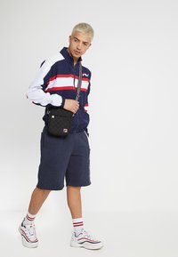 Giacca a vento blu navy con strisce rosse e bianche, abbinata a pantaloni corti blu navy. Borsa a tracolla nera, sneakers bianche con dettagli rossi.
