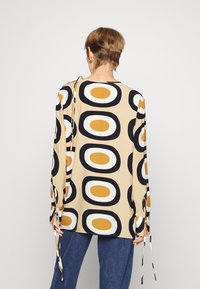 Blusa de manga longa em tecido leve, com fundo bege e grandes padrões ovais em preto, branco e mostarda; punhos atados.
