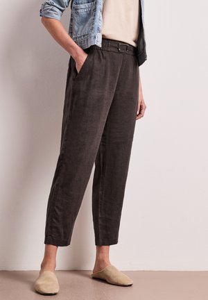 Persona che indossa pantaloni marrone scuro a vita alta con cintura, top beige, giacca di jeans risvoltata e scarpe beige slip-on, in piedi contro un muro chiaro.