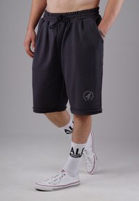 Schwarze Fleece-Shorts mit elastischem Bund und Kordelzug. Seitentaschen, umgeschlagene Säume und ein Logo-Patch am linken Bein. Weiße Turnschuhe und Socken.