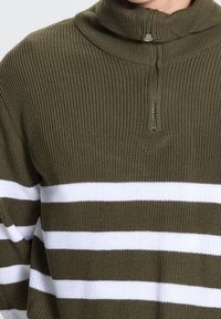 Primer plano de un suéter verde oliva acanalado con rayas blancas horizontales y un cuello alto parcialmente cerrado.