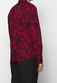 Langarmshirt van zwarte stof met een rode bloemenprint. Heeft een knoopsluiting en een ontspannen pasvorm, met een gebogen zoom.