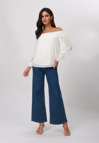White off-the-shoulder blouse with textured polka dots, featuring long puff sleeves. Paired with wide-leg blue denim jeans.