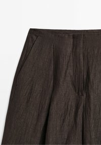 Gros plan d’un pantalon en lin brun foncé montrant la ceinture, les plis, la poche latérale et la braguette avant avec un tissu texturé.
