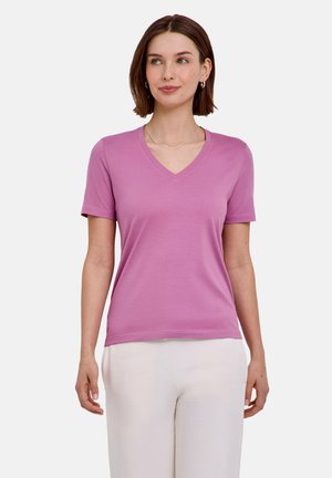 Vrouw met kort bruin haar die een roze T-shirt met V-hals en korte mouwen draagt en een witte broek, staand tegen een effen lichte achtergrond.