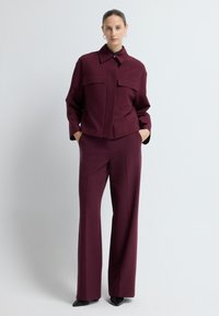 Bordeaux op maat gemaakte blazer met twee borstzakken en een open kraag, gecombineerd met bijpassende wijde broek. Gladde textuur en moderne snit.