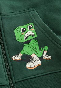 Grüner Hoodie mit einer Cartoonfigur mit grünem, kastenförmigem Kopf und ausdrucksstarkem Gesicht, die weiße Schuhe und rot gestreifte Socken trägt.