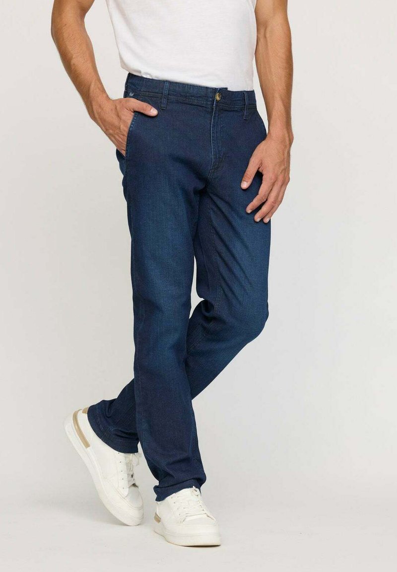 Jean en denim bleu foncé à coupe droite, avec des poches avant et une fermeture à bouton, portés avec des baskets blanches sur un fond neutre.