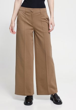 Femme portant un pantalon marron taille haute à jambes larges, associé à des mocassins noirs en cuir tissé et un haut noir sur un fond uni.