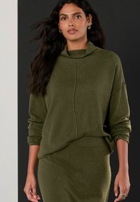 Zielony dzianinowy sweter z wysokim kołnierzem, prążkowanymi mankietami i dołem. Gładka faktura, luźny krój, zestawiony z pasującą spódnicą.