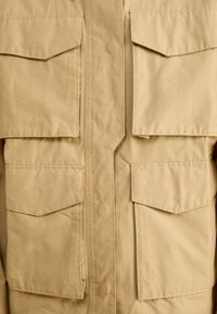 Beżowa kurtka z tkaniny z przednim zamkiem błyskawicznym, czterema dużymi kieszeniami z klapami oraz konstrukcją z widocznymi detalami szwów.