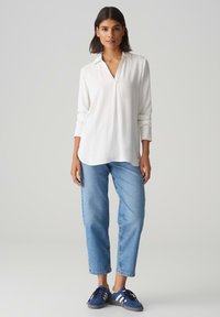 Weiße langärmelige Bluse mit V-Ausschnitt und Kragen, kombiniert mit hellblauen Jeans und dunkelblauen Sneakers mit weißen Streifen.