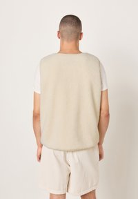 Vyriškis apsirengęs kreminės spalvos flišine liemene virš baltos trumpomis rankovėmis palaidinės ir šviesių kordurojų šortų, stovintis atsisukęs nugara priešais paprastą foną.