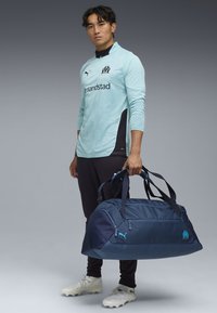Sac de sport en toile marine avec plusieurs poches zippées, sangles en tissu et logo Puma. Surface texturée avec une finition lisse, notamment tenu par un mannequin.