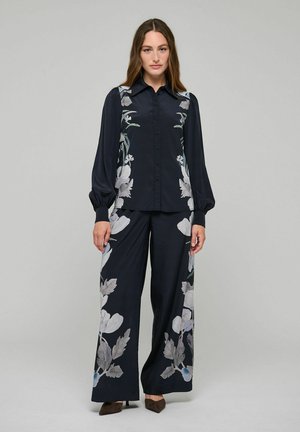 Donna in piedi che indossa una blusa nera con bottoni e pantaloni a gamba larga abbinati con grandi stampe floreali bianche e grigie.