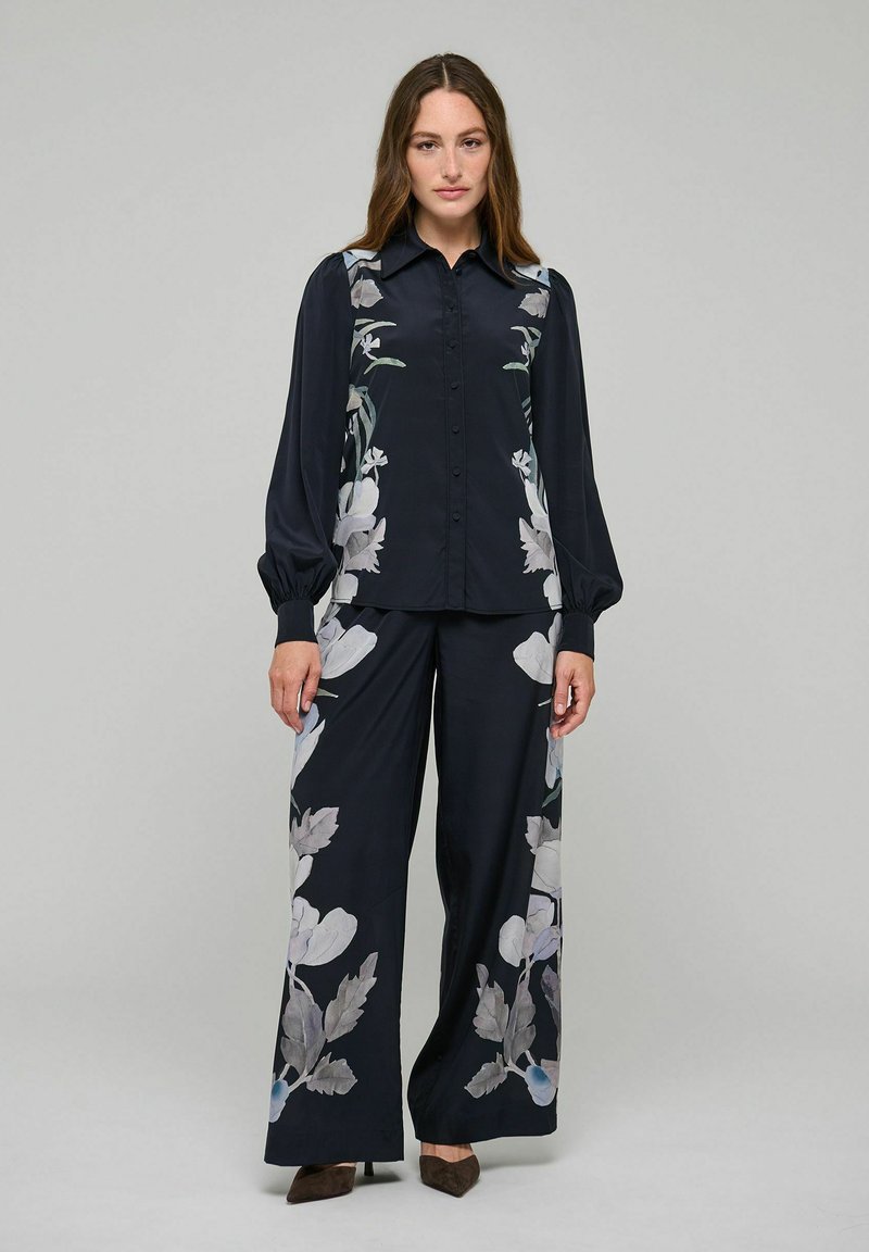 Femme debout portant un chemisier noir boutonné et un pantalon large assorti avec de grands motifs floraux blancs et gris.